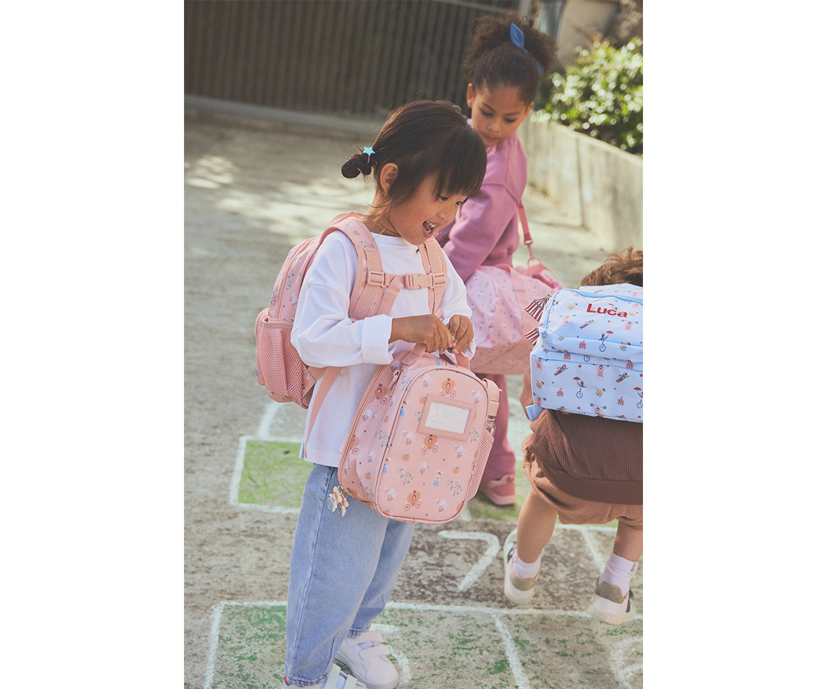 Crianças com mochilas coloridas e padrão infantil a brincar ao ar livre num chão de terra com amarelinha pintada.