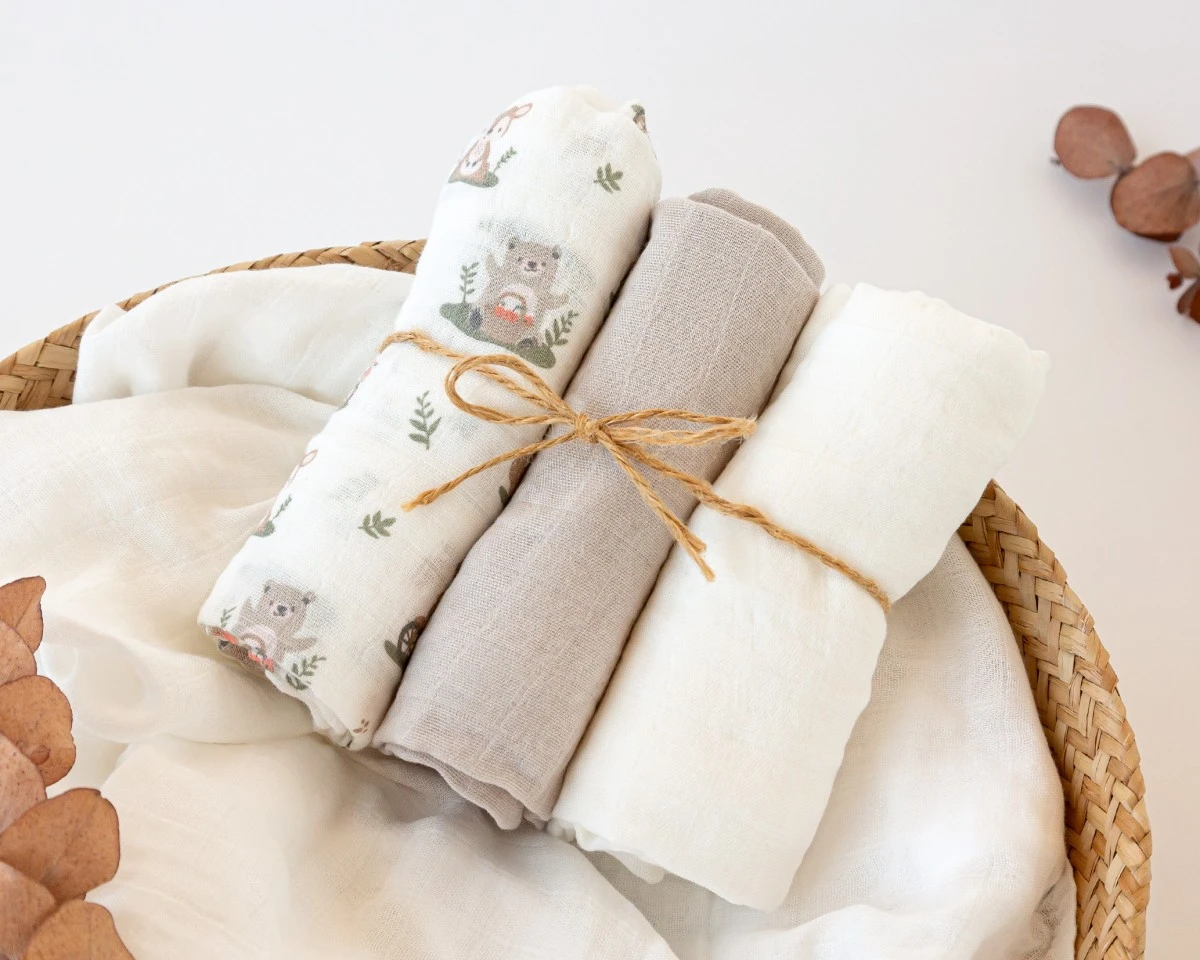 Três fraldas de algodão branco e cinzento com estampa de ursinhos, amarradas, em panier de vime sobre fundo branco