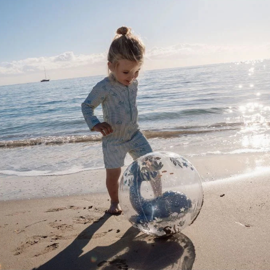 Criança com fato de banho azul claro às riscas na praia a brincar com bola transparente decorada com desenhos marinhos.