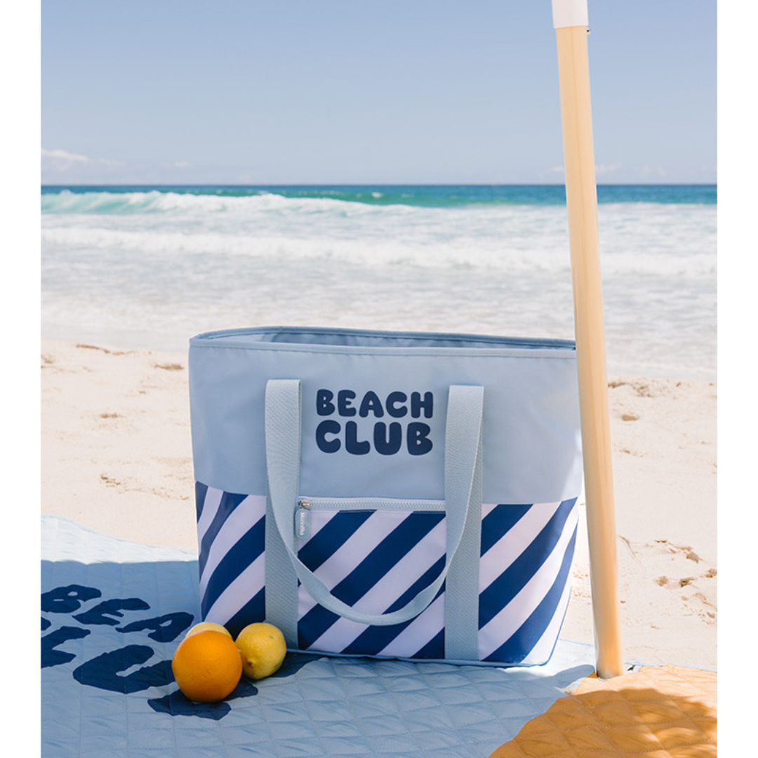 Saco de praia azul com riscas e texto BEATCH CLUB na areia junto a frutas e guarda-sol.