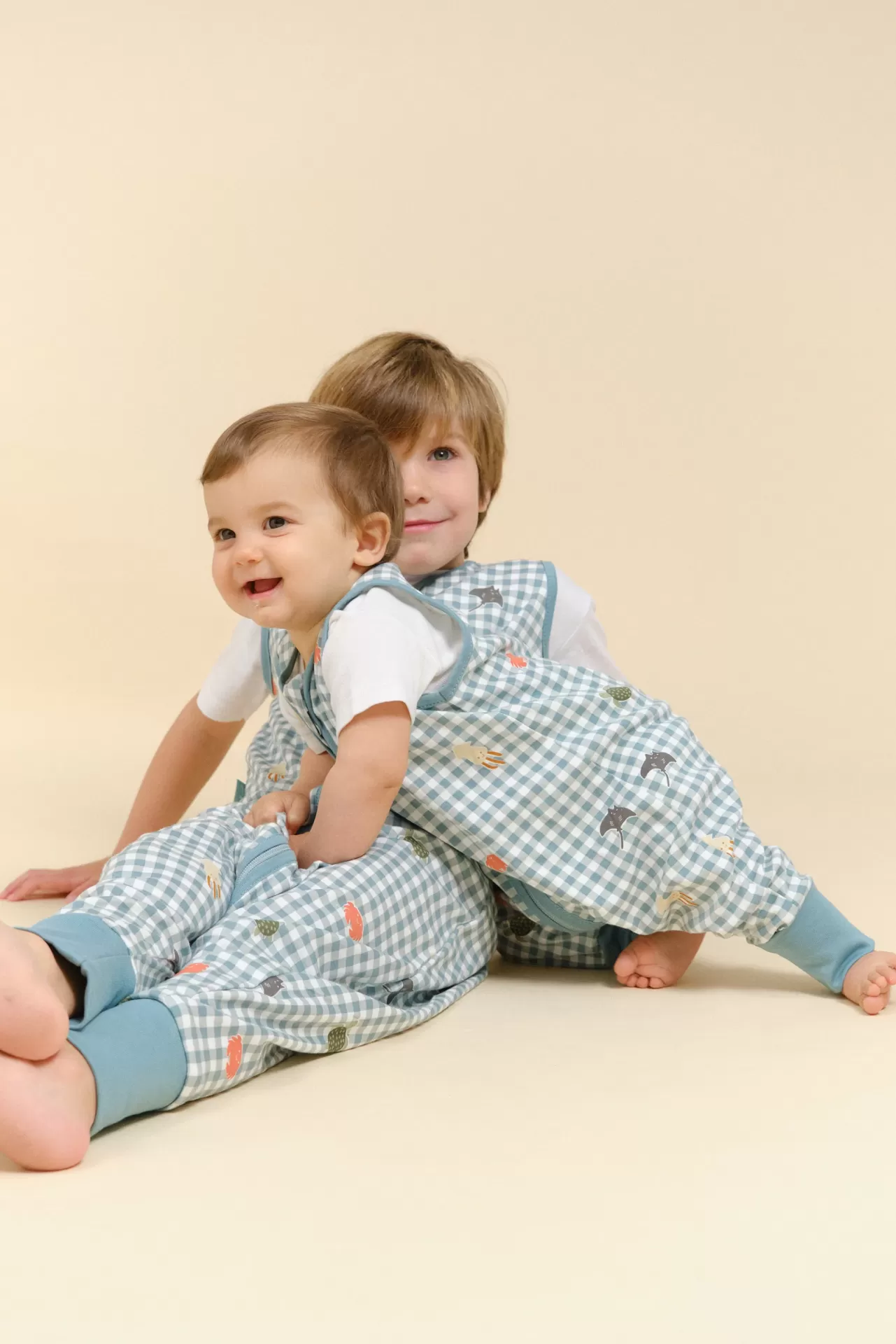 Duas crianças com macacões xadrez azul e branco estampados e fundo bege claro