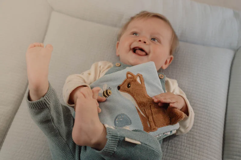 Bebé sorridente com jardineiras e livro tecido azul com raposa