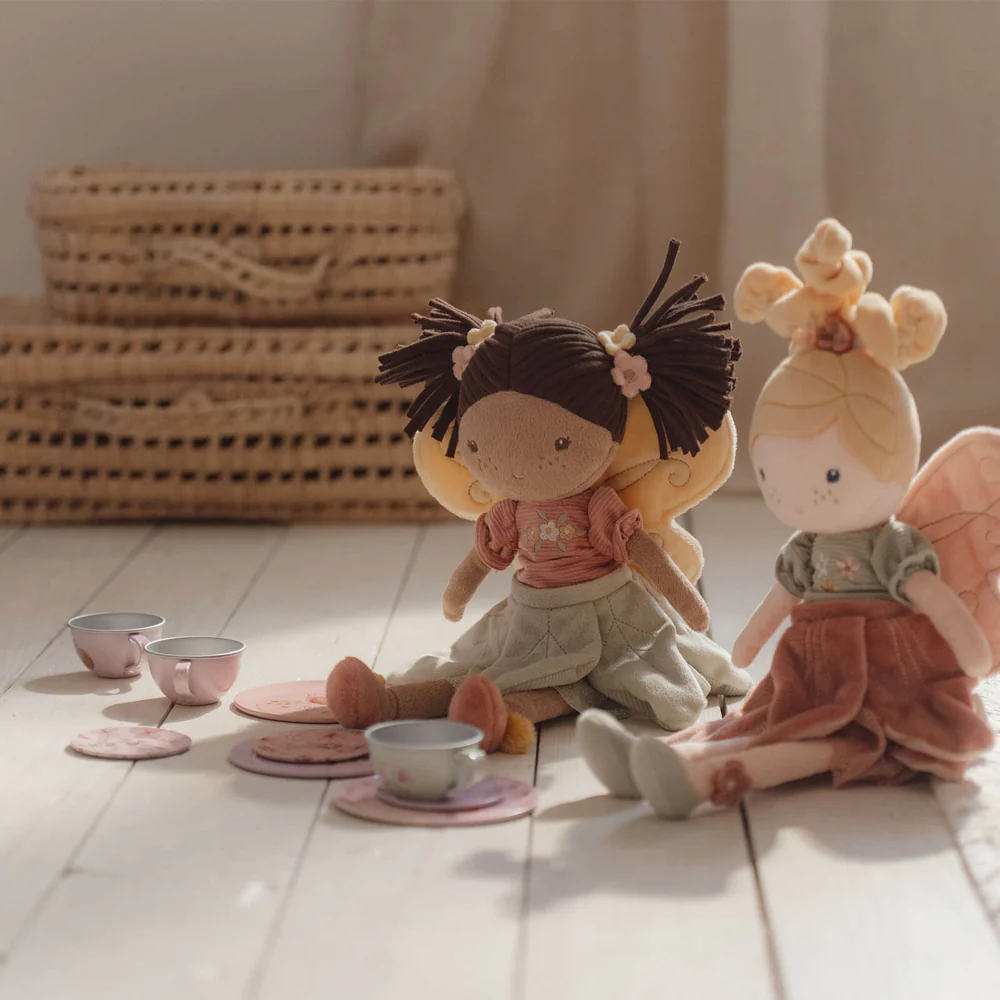Duas bonecas de pano com roupas de tecido sentadas perto de um conjunto de chá de brinquedo, com cestos de vime ao fundo