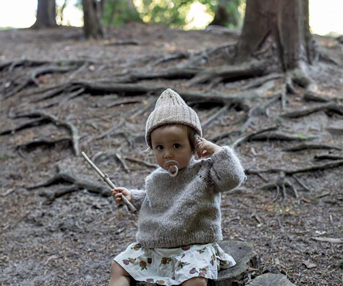 Criança com roupa de malha e chupeta sentada em tronco numa floresta