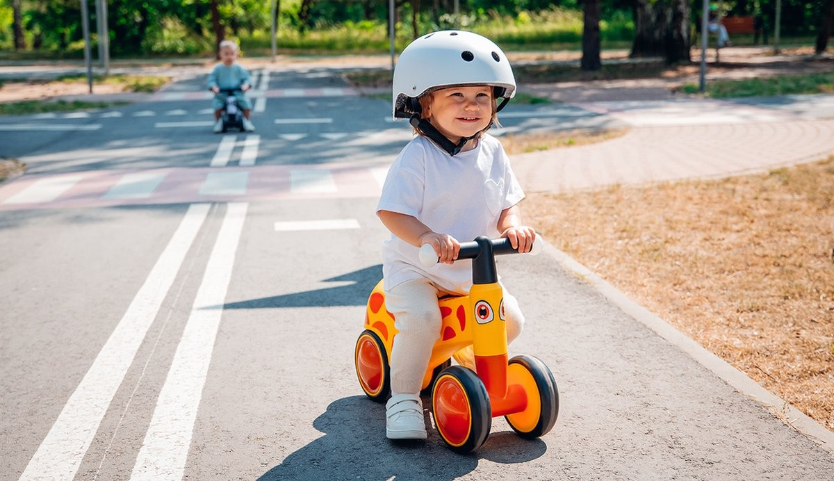 Criança com capacete branco num veículo de brincar amarelo e laranja numa estrada asfaltada com marcas e passadeira