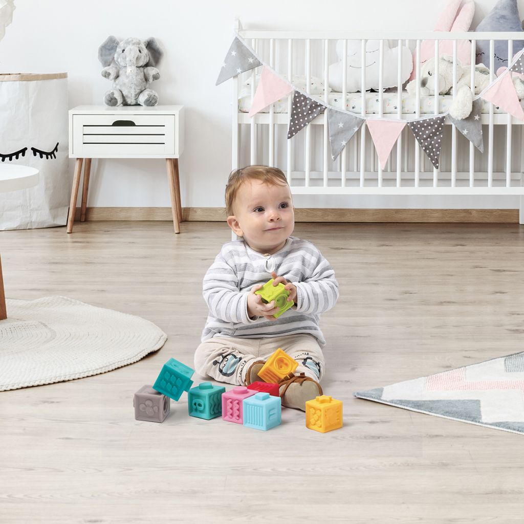 Bebé sentado a brincar com blocos coloridos num quarto infantil com berço branco e decoração em tons pastel.