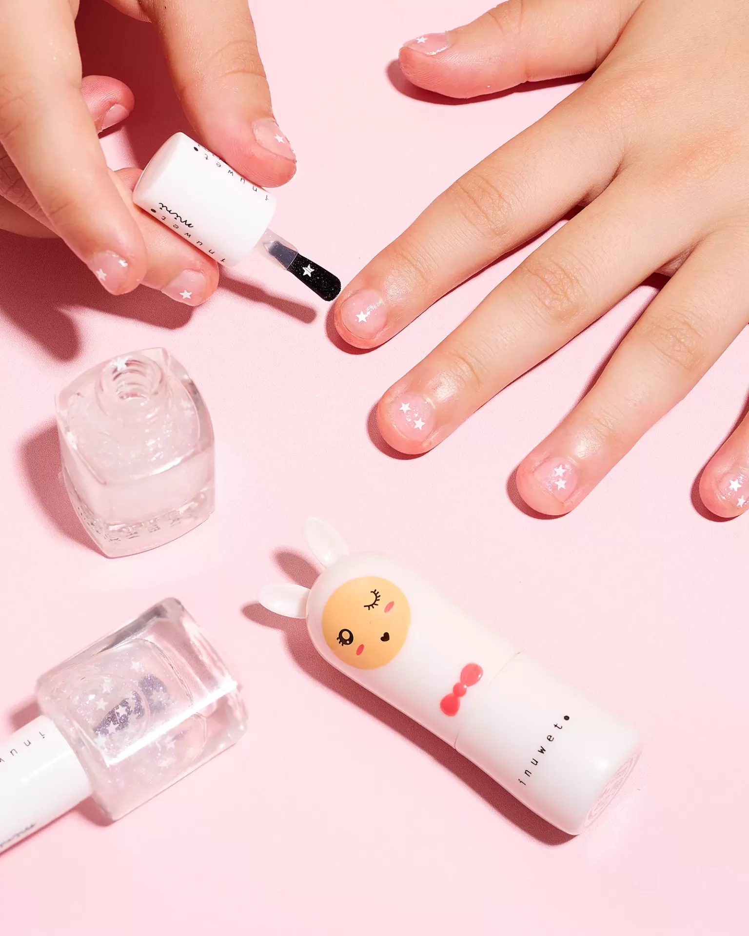 Mãos de criança a aplicar verniz transparente com estrelas e produtos de verniz sobre fundo rosa.