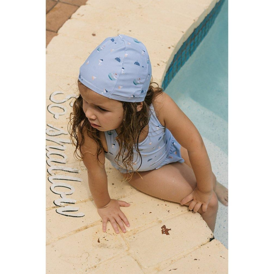 Criança com fato de banho azul claro e touca de banho junto a piscina