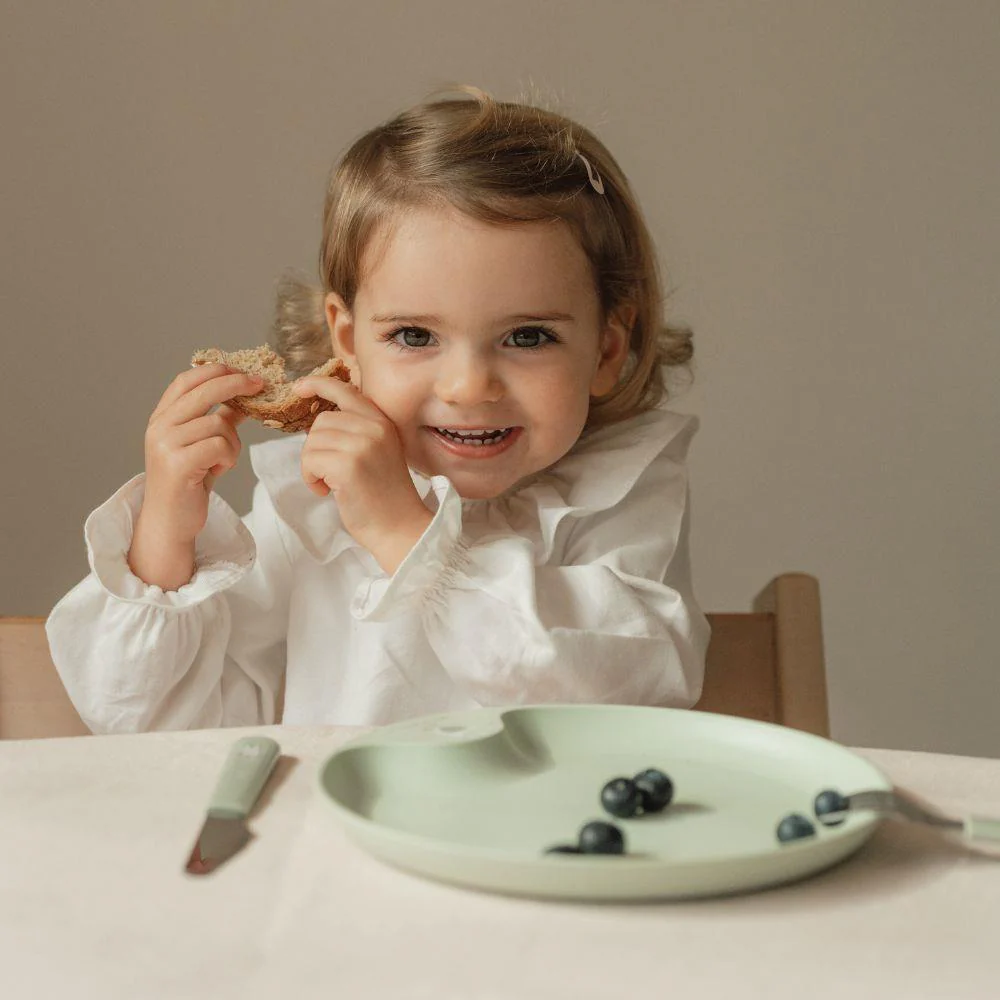 Criança sorridente segurando pão à mesa com prato e mirtilos