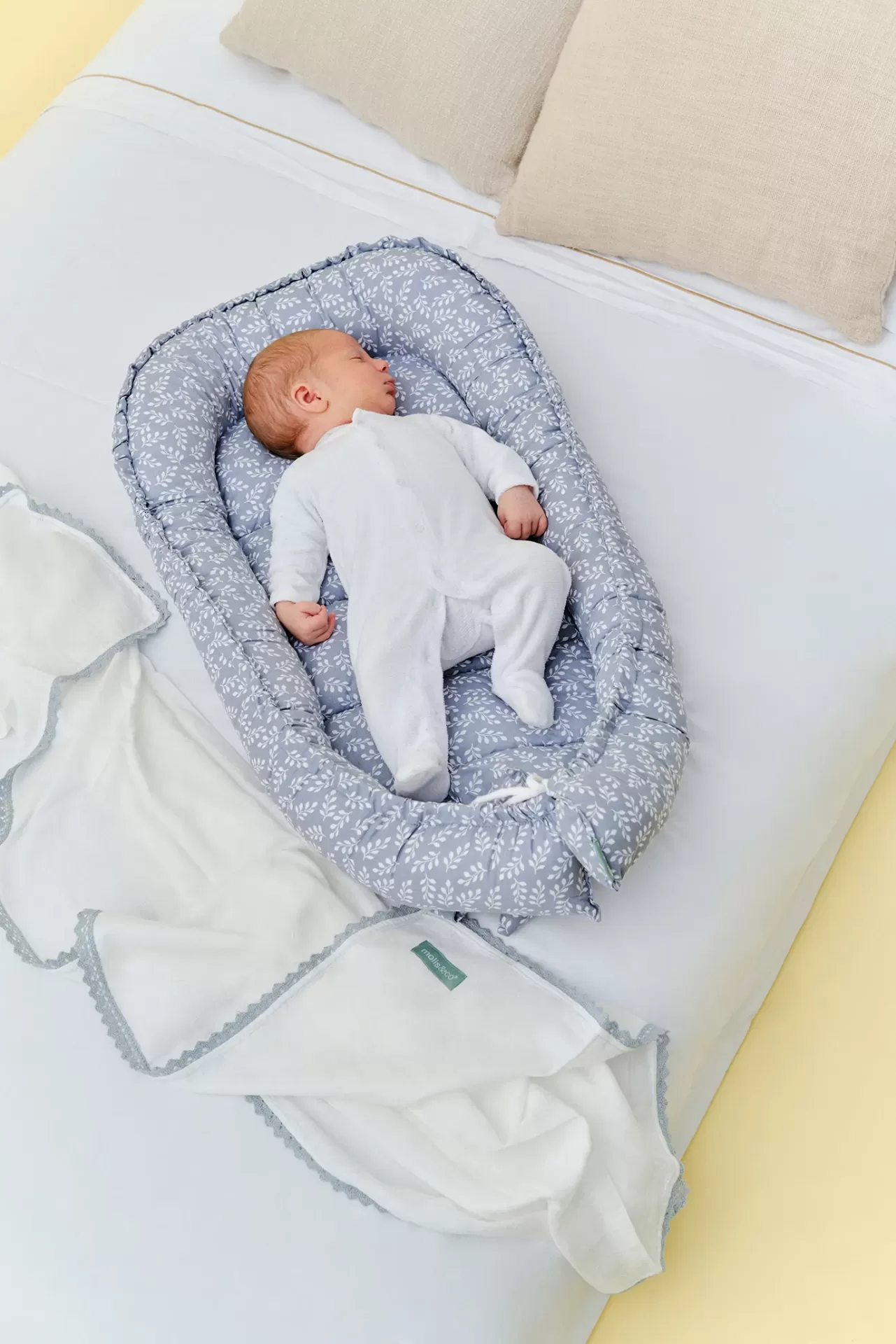 Ninho de bebé cinzento com padrão branco, cobertor branco com borda cinzenta e bebé de branco a dormir numa cama