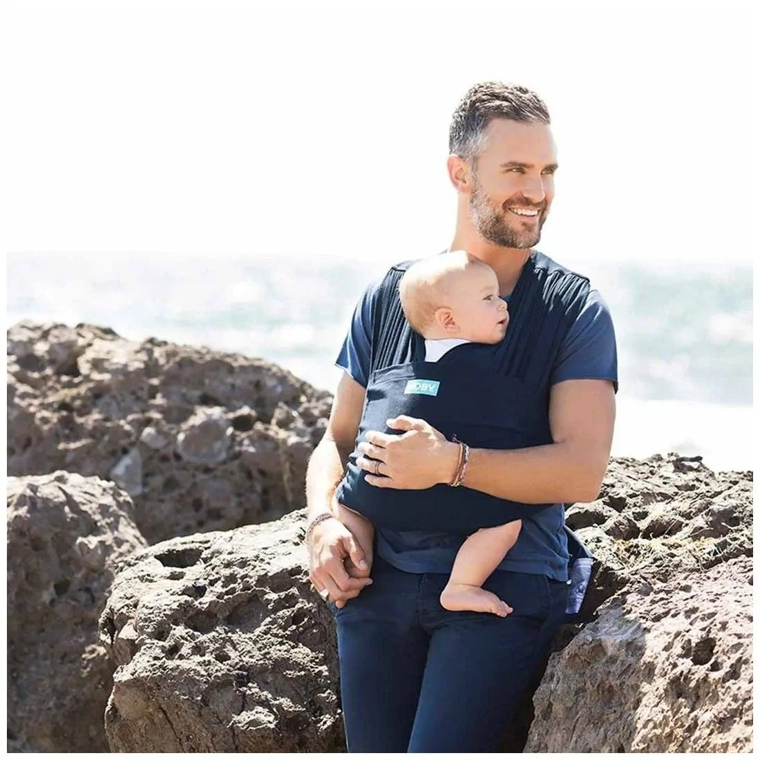 Homem com bebé em canguru azul escuro sentado em rochas perto do mar