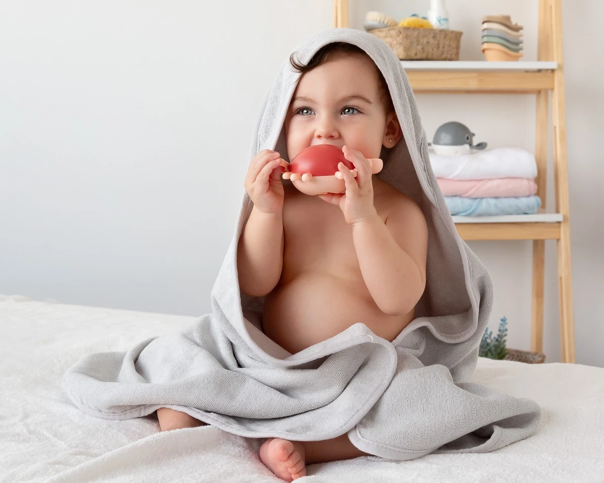 Bebé com toalha cinza segurando brinquedo vermelho e bege sentado em cama branca