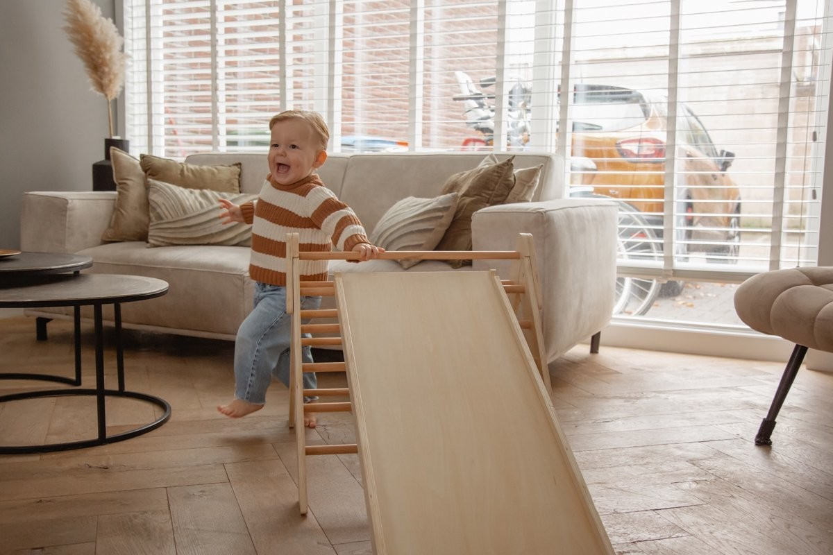 Criança a brincar numa rampa de madeira numa sala com sofá bege e almofadas castanhas