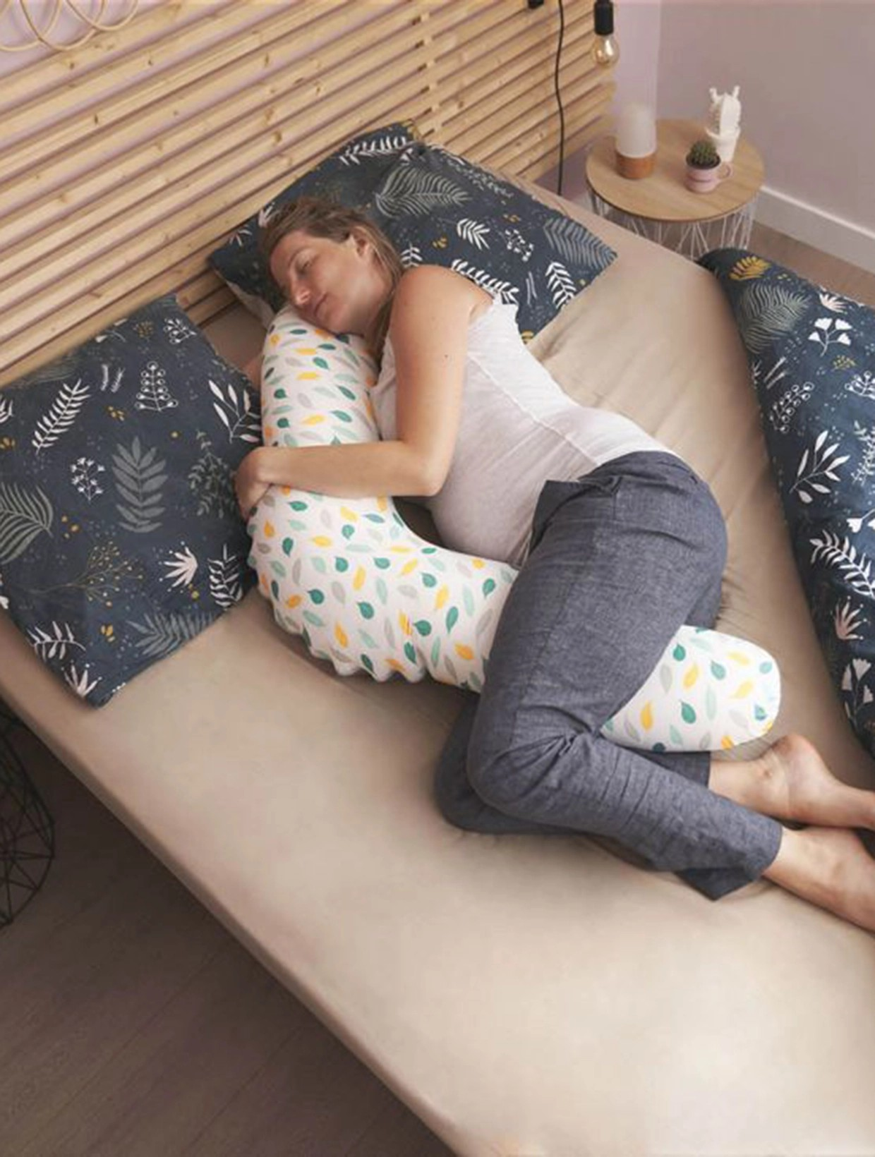 Mulher grávida a dormir e abraçada a almofada de gravidez com padrão de folhas coloridas numa cama com almofadas azuis e lençóis bege