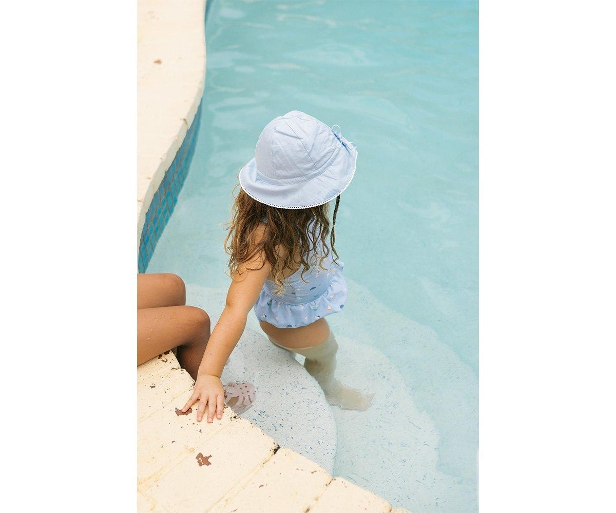 Criança de chapéu branco e maiô branco na borda de piscina com água azul