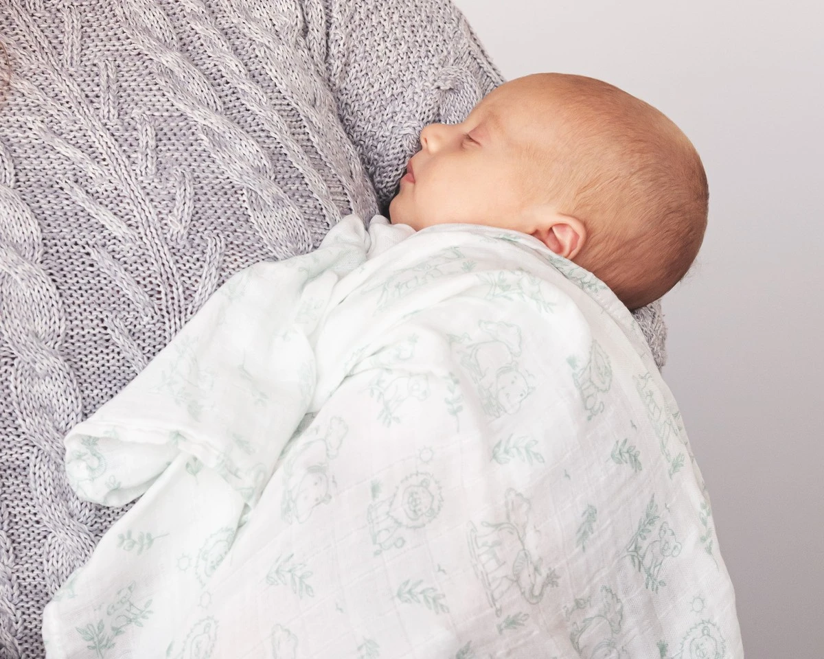 Bebé adormecido enrolado numa manta branca com padrões em verde claro apoiado no peito de uma pessoa com camisola cinzenta de malha.