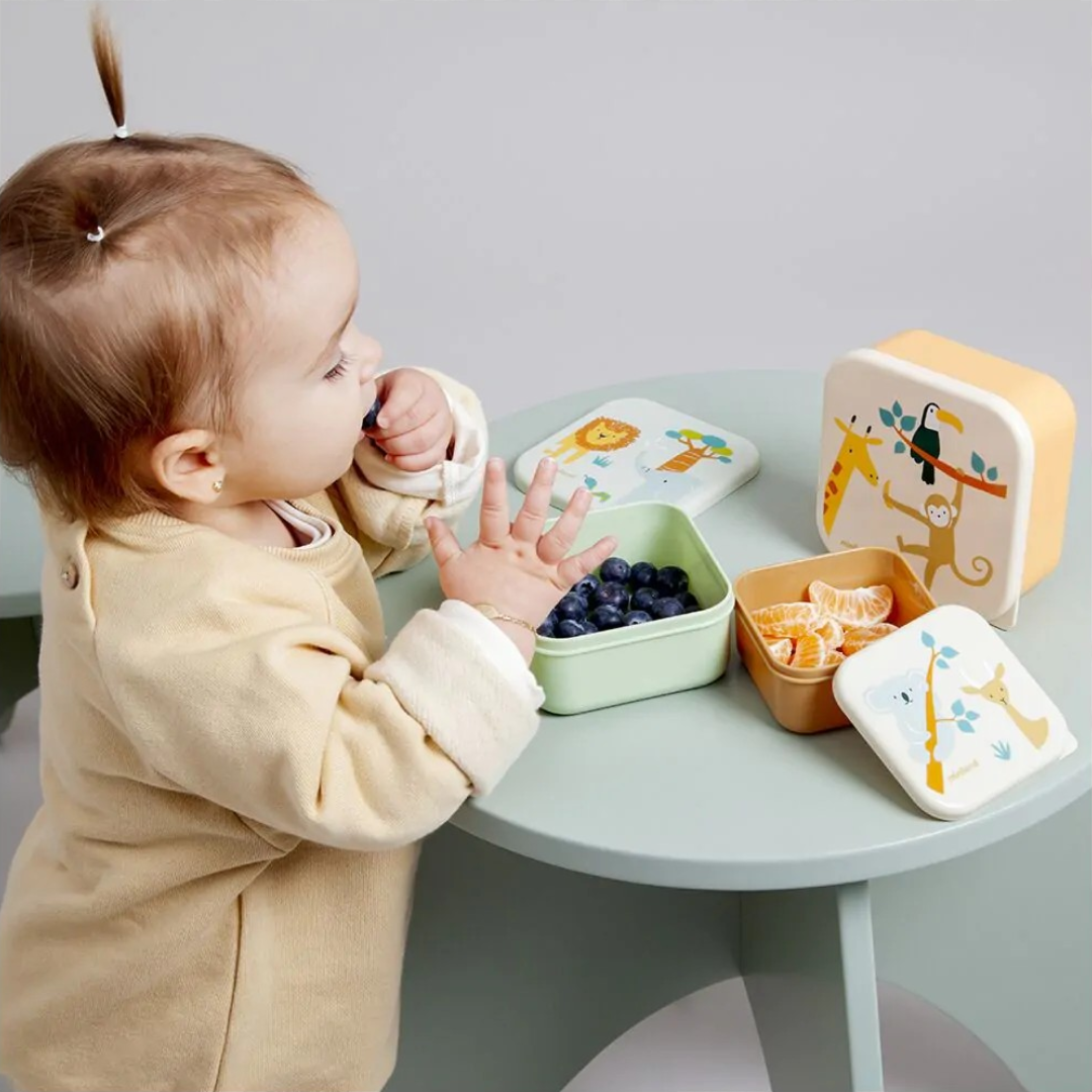 Bebé a comer frutas em recipientes de plástico com tampas decoradas com animais sobre mesa verde-água