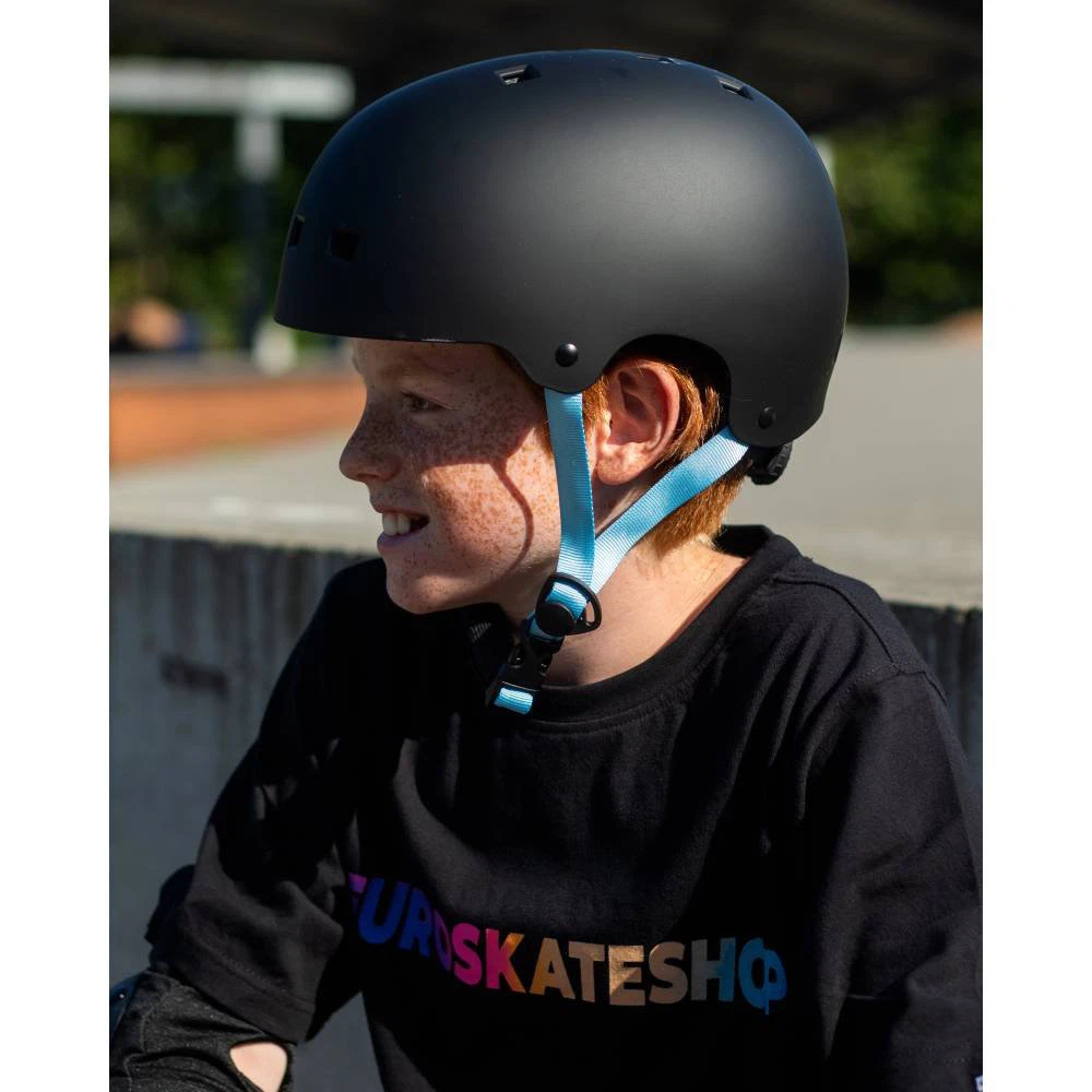Criança com capacete preto mate e t-shirt preta com texto colorido EUROSKATESHOP sentada no parque de skate.
