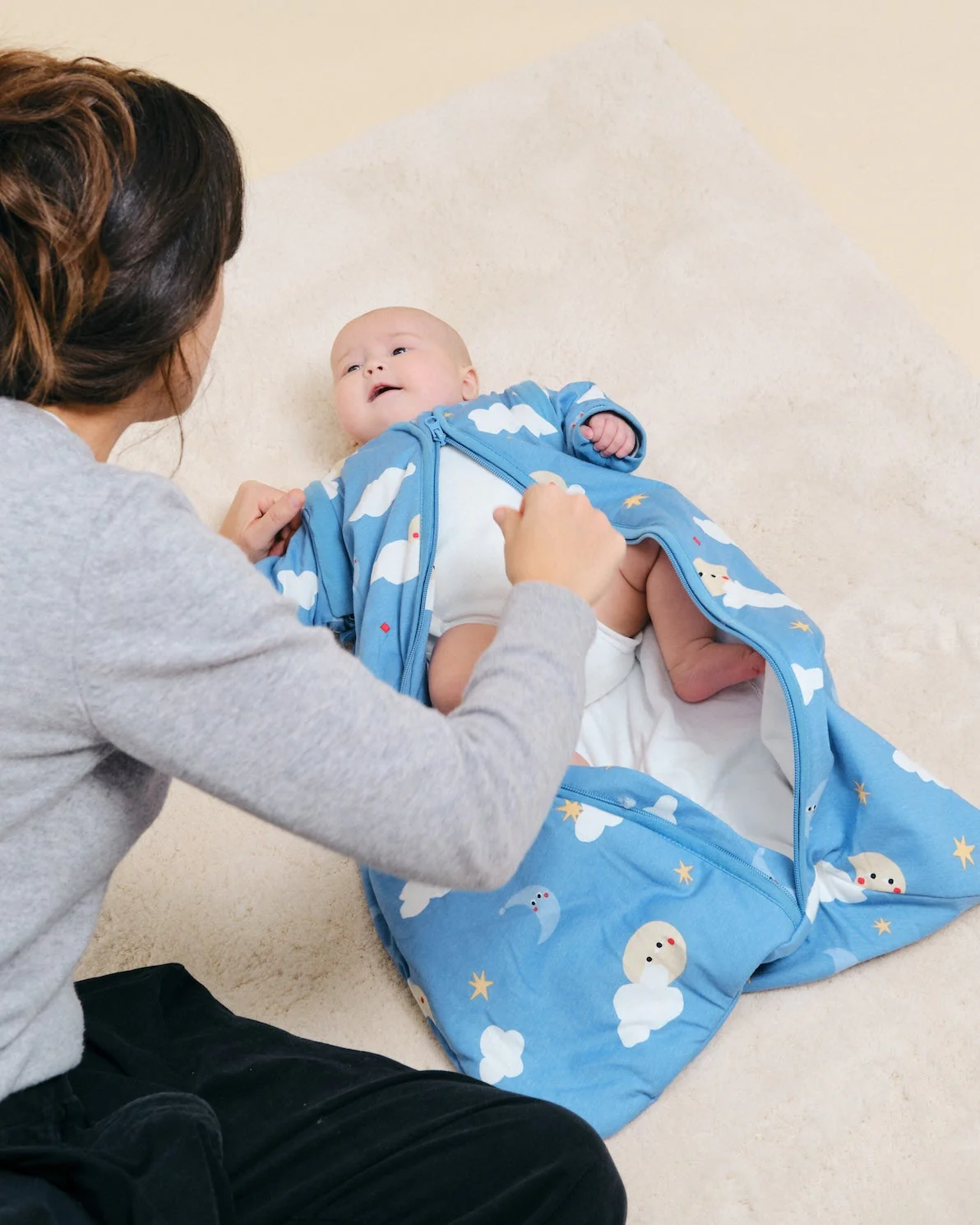 Saco de dormir azul com padrão de nuvens, luas e estrelas para bebé num tapete creme.