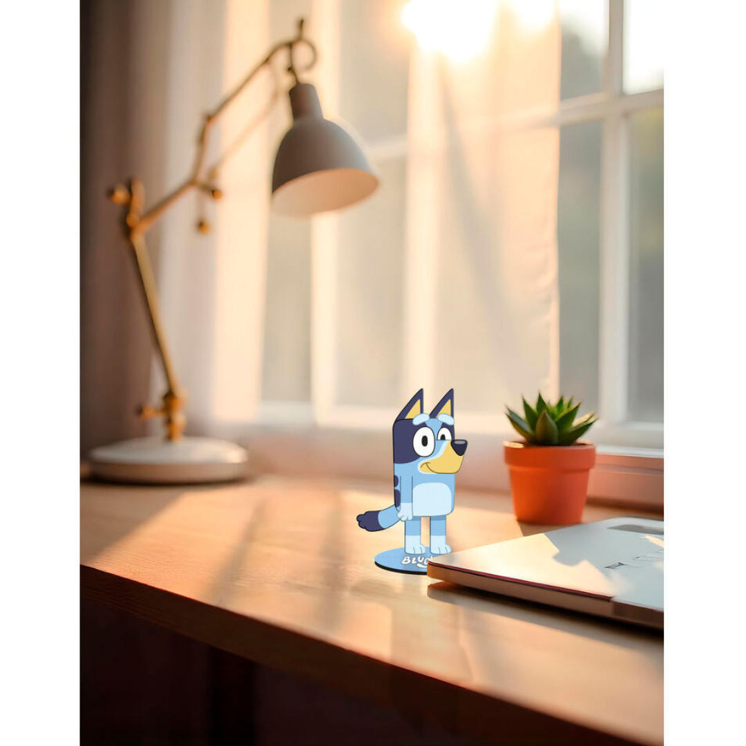 Figura de papel do personagem Bluey sobre mesa com portátil, planta e lâmpada