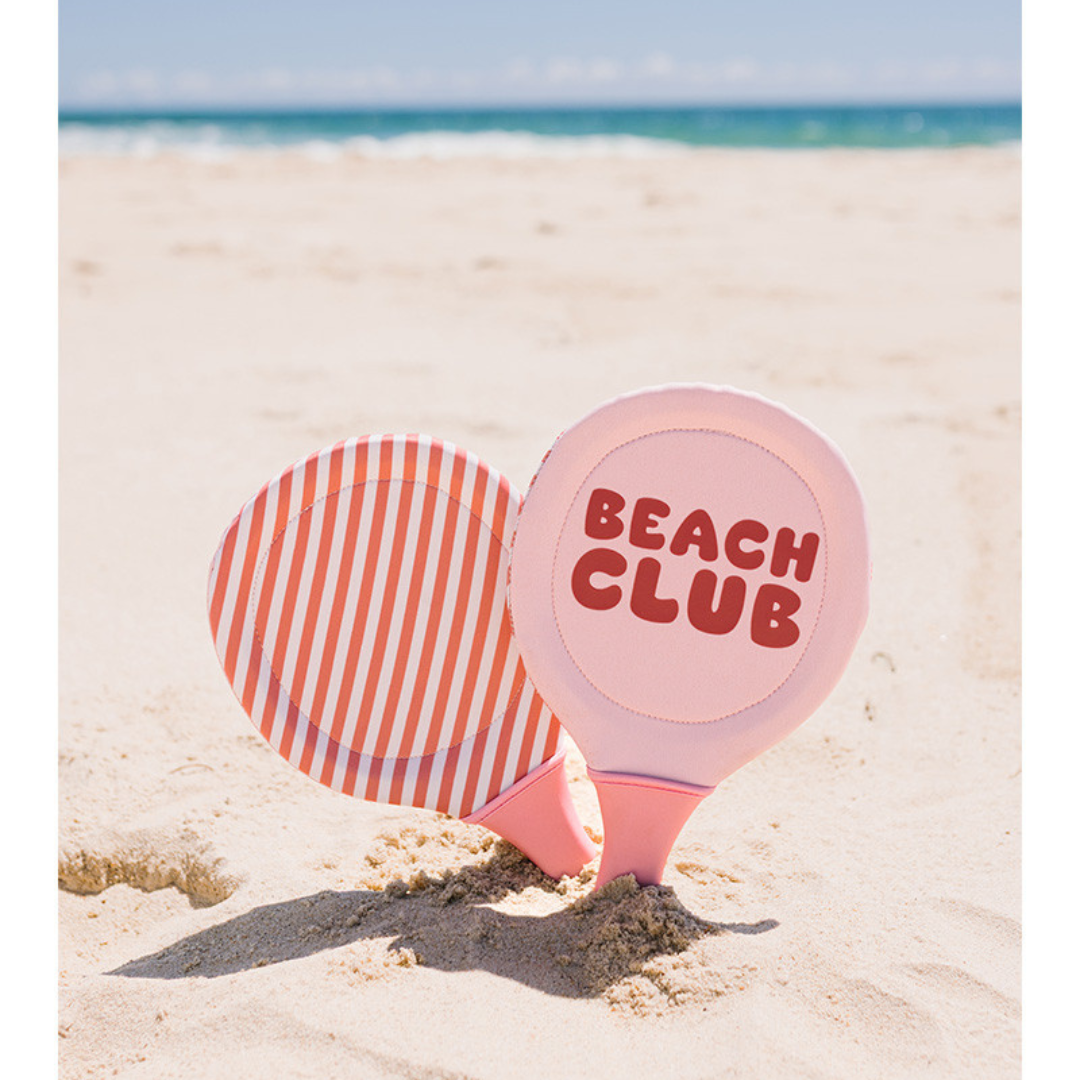 Raquetes de praia rosas com padrão riscado e texto BEACH CLUB na areia junto ao mar