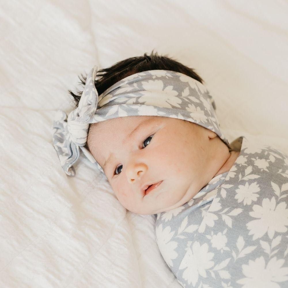 Bebé com faixa de cabelo e envoltório floral cinza claro deitado em tecido branco