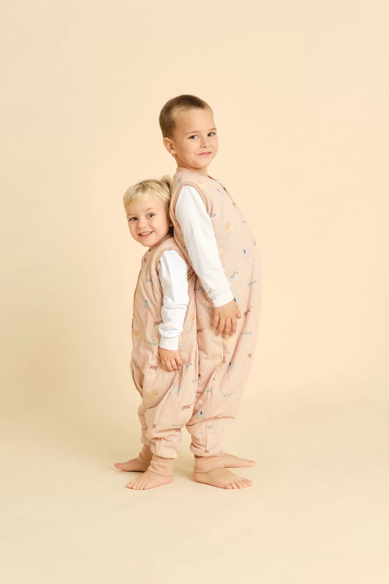 Dois rapazes em fatos cor de pêssego com desenhos de animais e natureza, ao pé descalços sobre fundo bege.