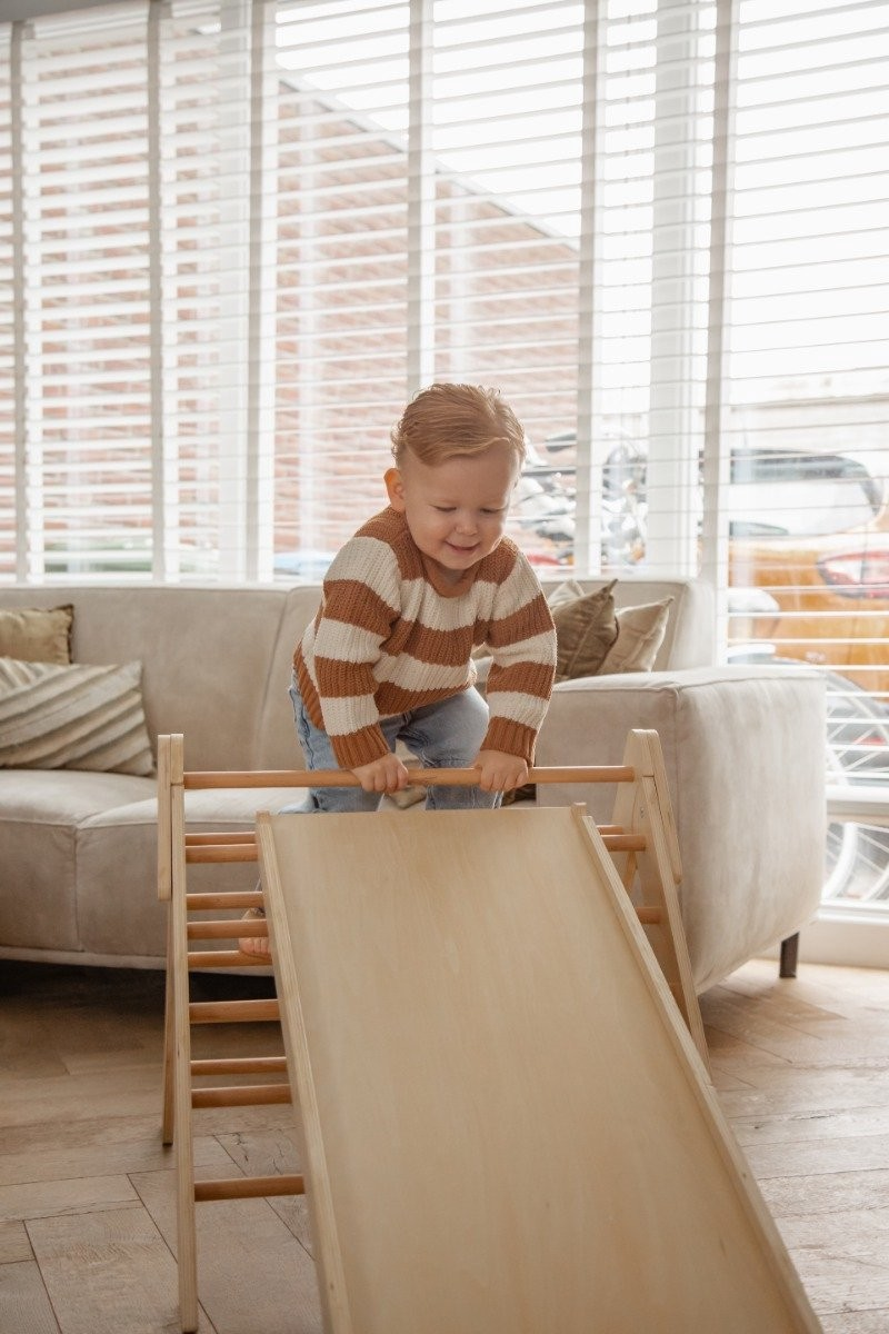 Criança a brincar num escorrega de madeira em sala com sofá e persianas
