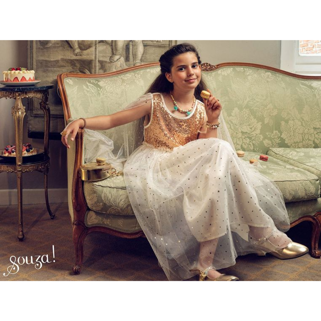 Menina com vestido branco e dourado sentada num sofá verde com macarons e tortas ao lado