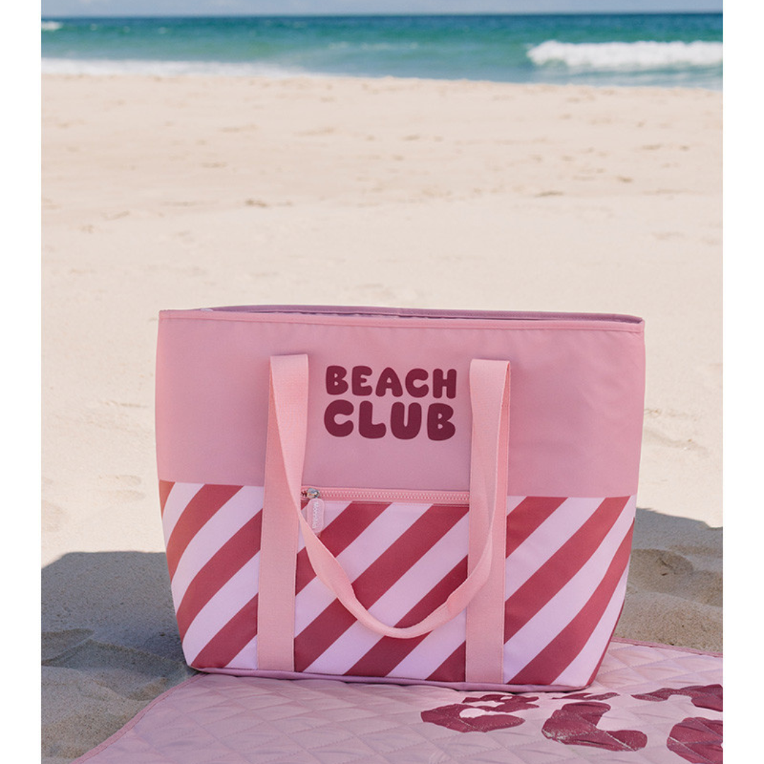 Sacola de praia rosa com padrão diagonal e texto Beach Club sobre toalha de praia na areia