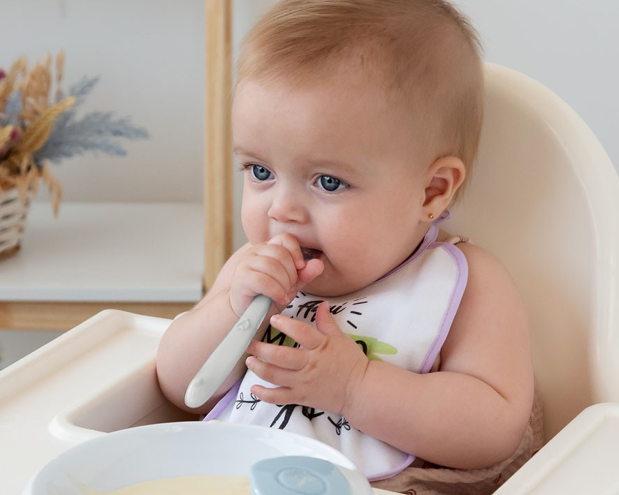 Bebé sentada numa cadeira de alimentação a segurar uma colher e com babete