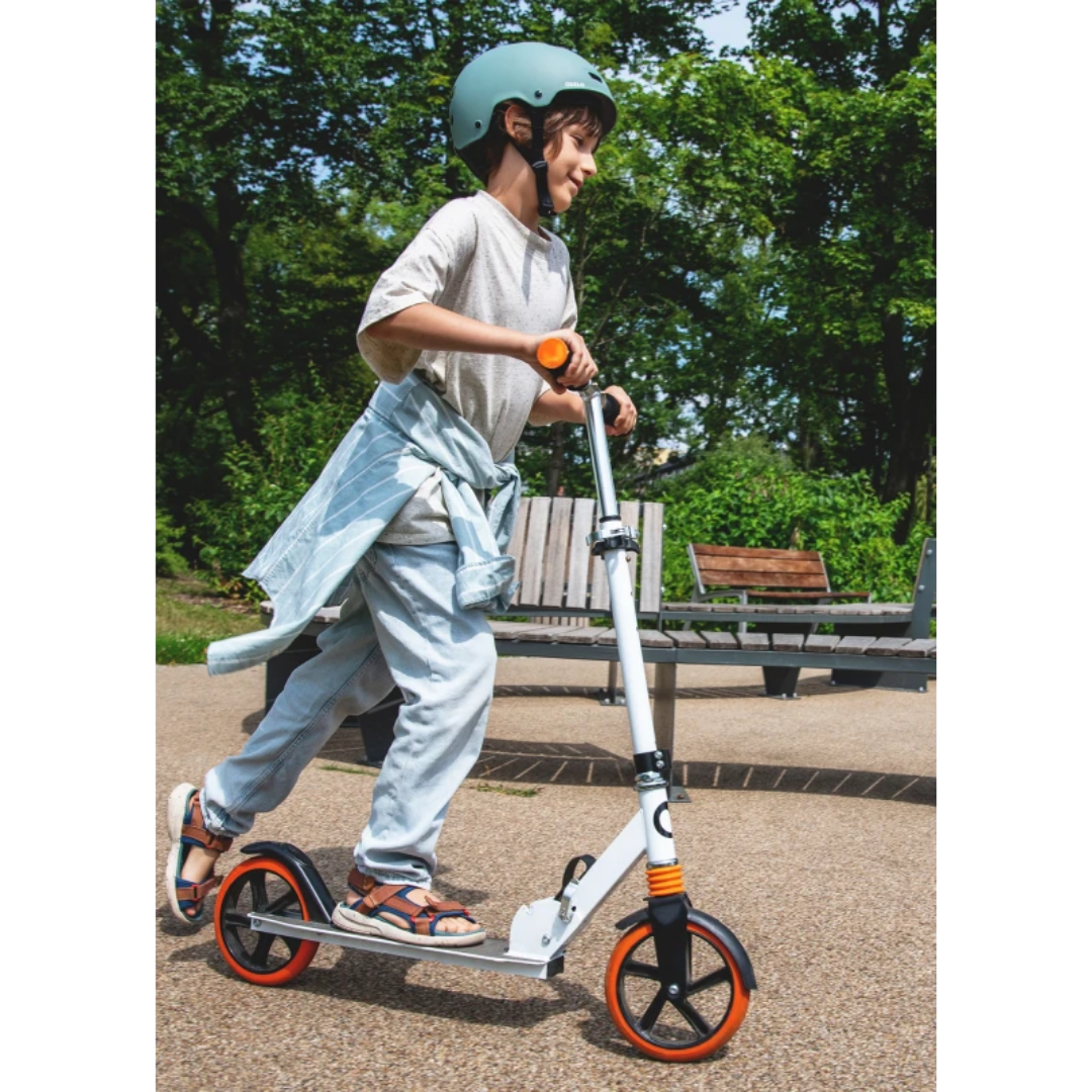 Criança a andar de trotinete branca com rodas laranja num parque com bancos e árvores.