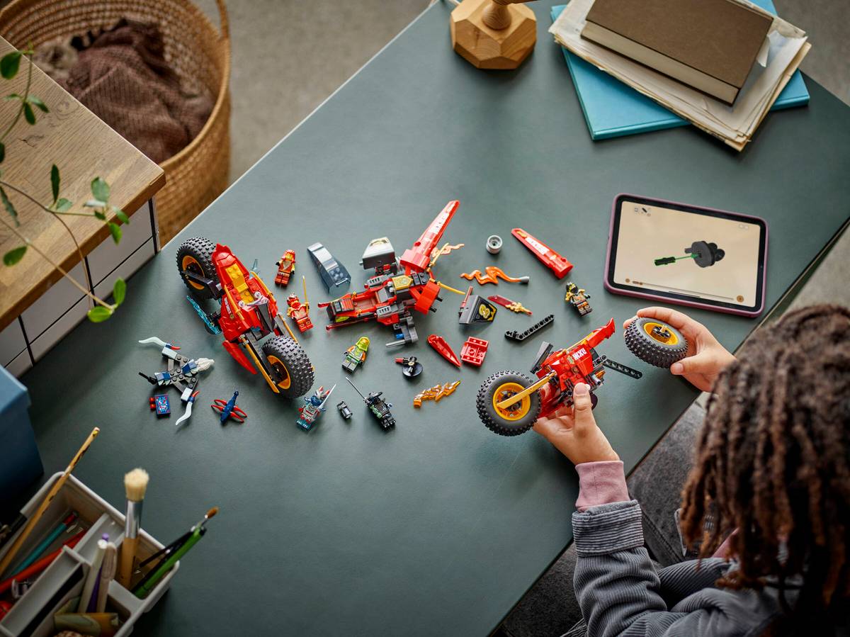 Peças de construção tipo Lego com minifiguras e scooters vermelhas numa mesa cinza com criança a montar.