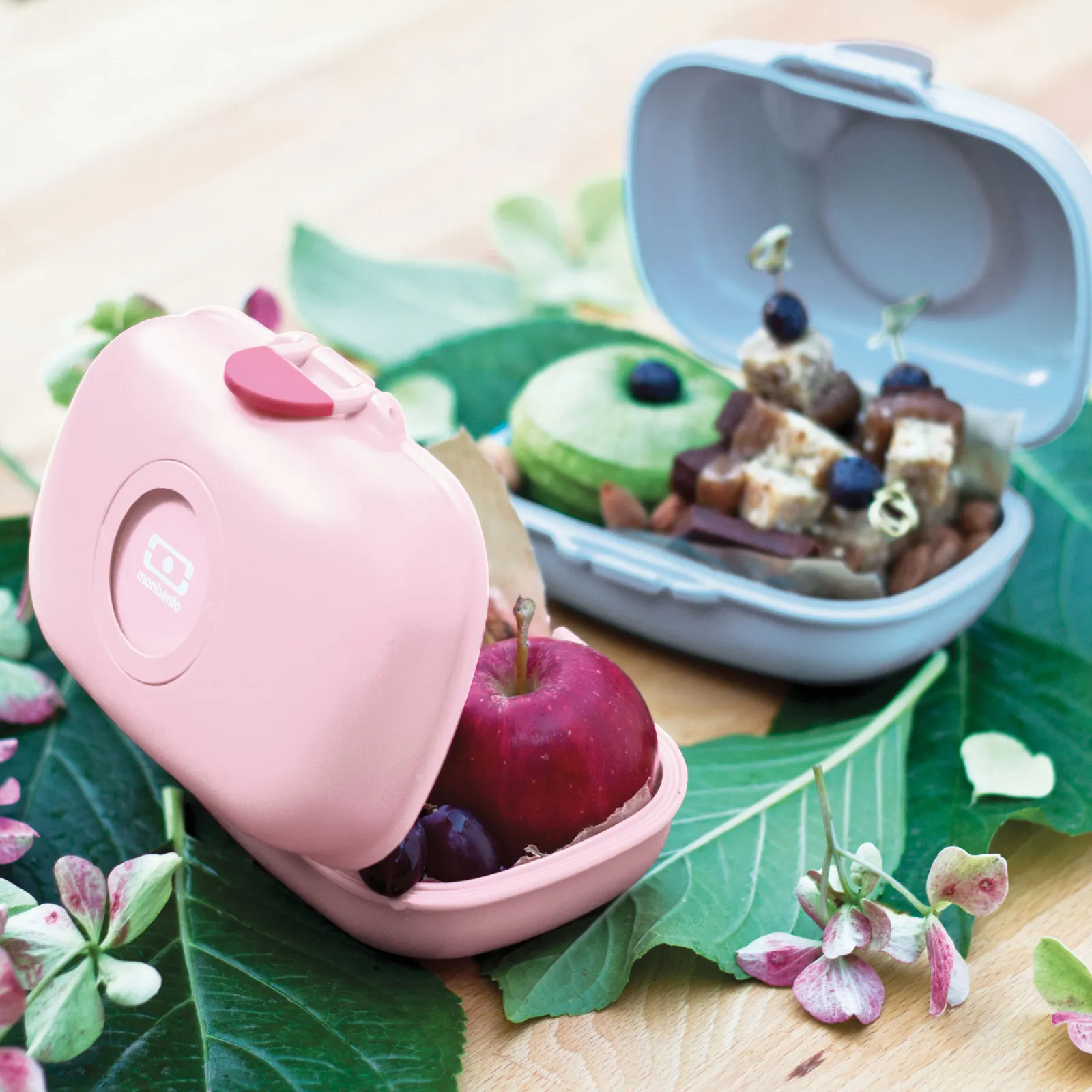 Caixas de lanche rosa e cinza com frutas e snacks sobre folhas e flores