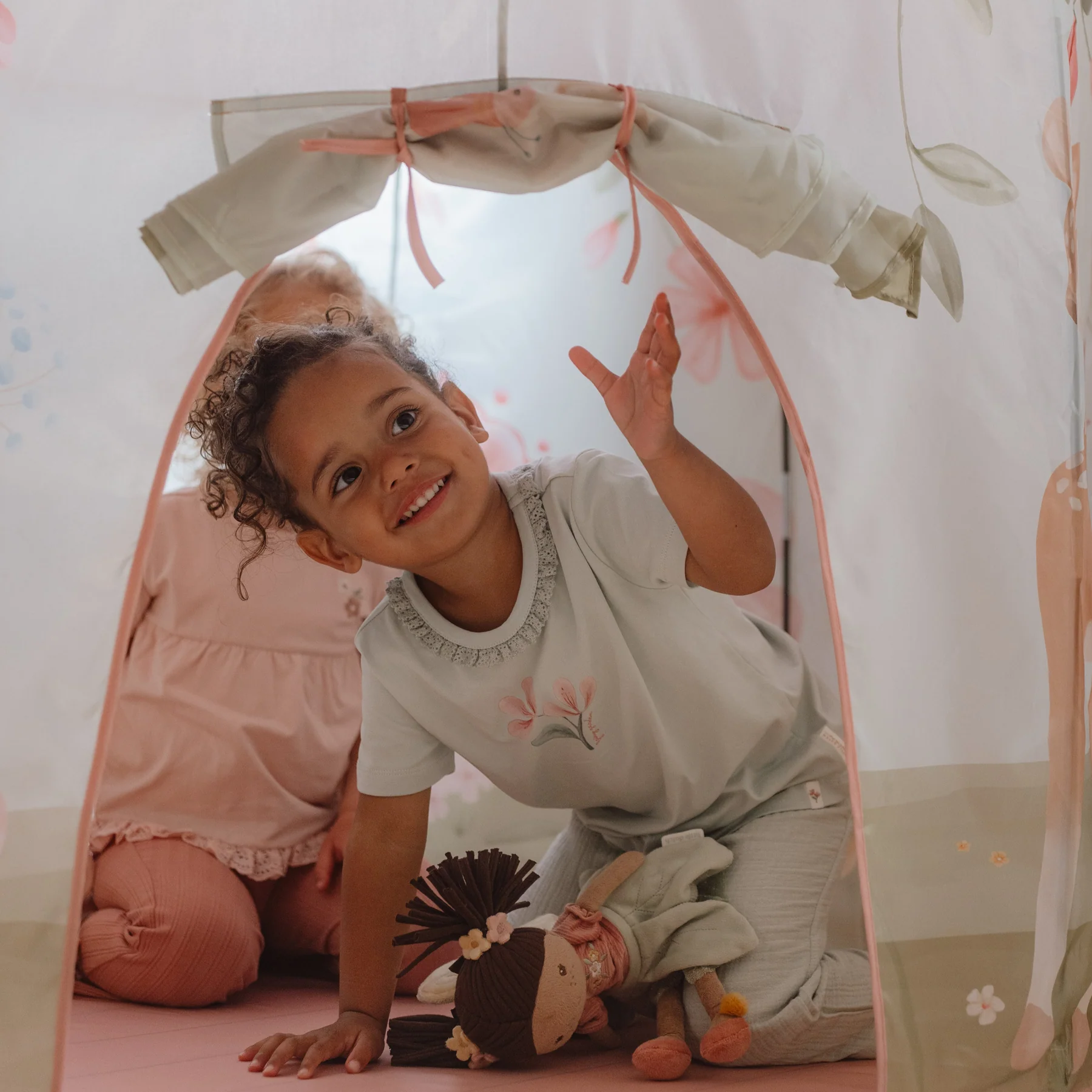 Criança sorridente dentro de tenda de tecido com padrão floral, vestindo pijama cinza com motivo floral e segurando uma boneca de pano
