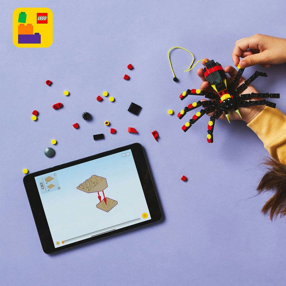 Mão segurando aranha preta, vermelha e amarela feita com peças LEGO ao lado de tablet com instruções e peças LEGO soltas