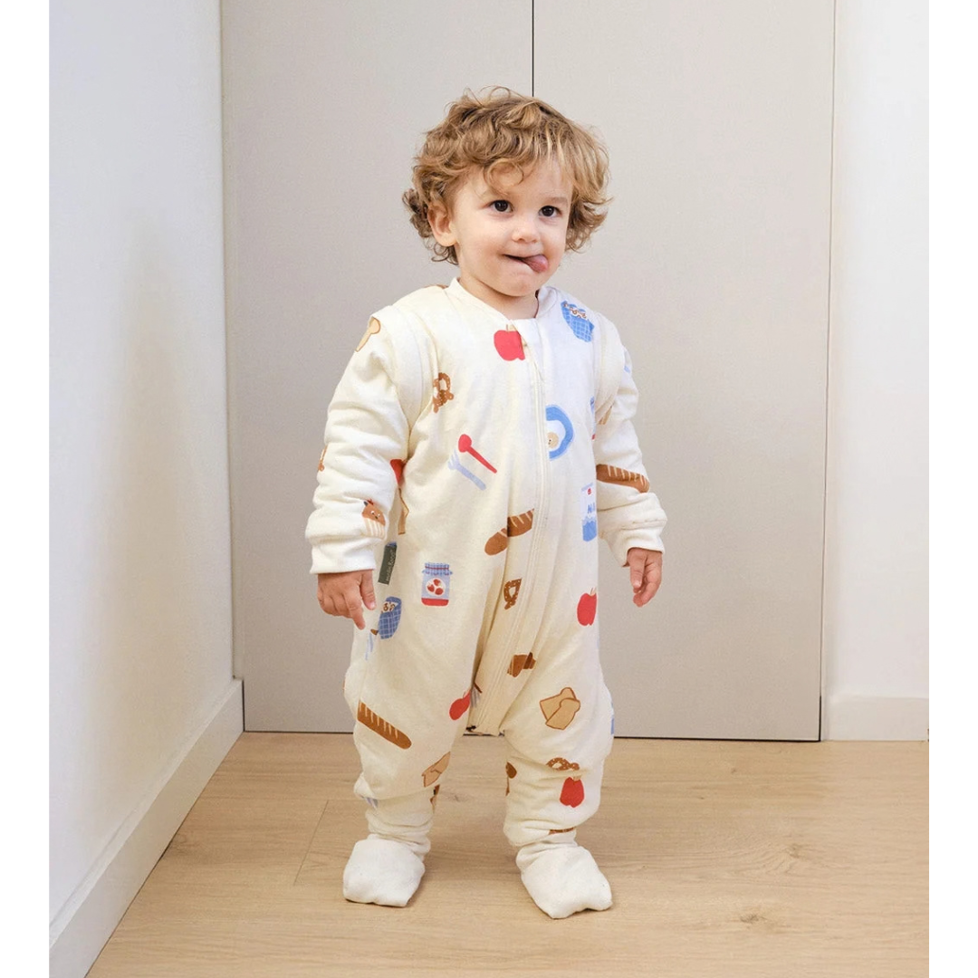 Criança com macacão creme estampado com motivos de pão, maçãs e potes, fecho frontal zipper, meias brancas, em chão madeira clara e parede branca