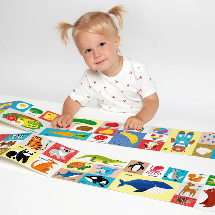 Criança com t-shirt branca, olhando para livro/cartaz ilustrado com animais e alimentos coloridos sobre mesa branca