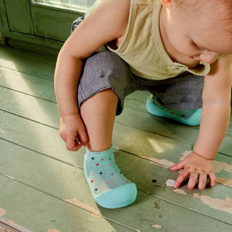 Criança com meias cor-de-menta e calções cinzentos a engatinhar no chão de madeira verde