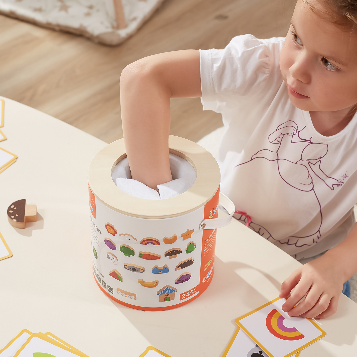 Brinquedo de jogo táctil cilíndrico com tampa de madeira e criança a jogar cartas coloridas.