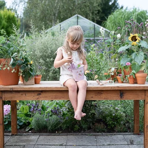 Criança com almofariz e pistilo sentada numa mesa de madeira rodeada por vasos com plantas num jardim