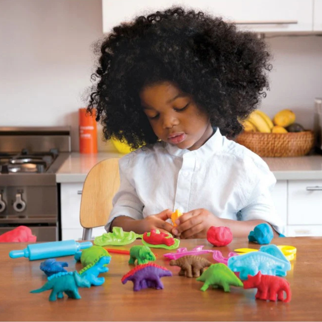 Criança a brincar com plastilina e pequenos moldes de dinossauros coloridos numa mesa de madeira na cozinha.