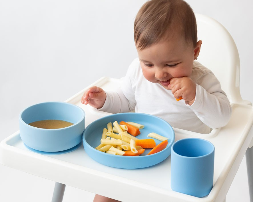 Bebé a comer numa cadeira alta com prato e copo azul.