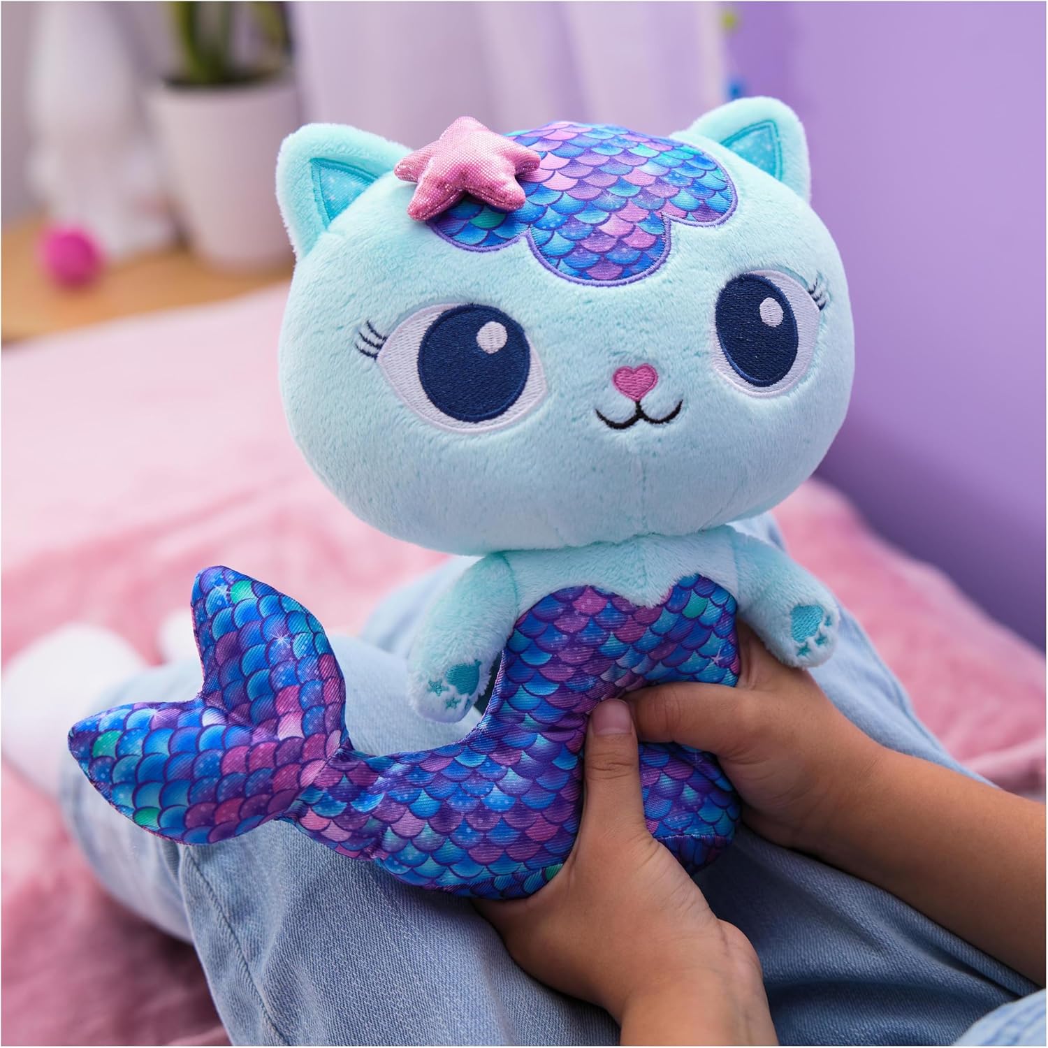 Pelúcia de gato sereia azul claro com escamas roxas e azuis, segurada por uma criança.