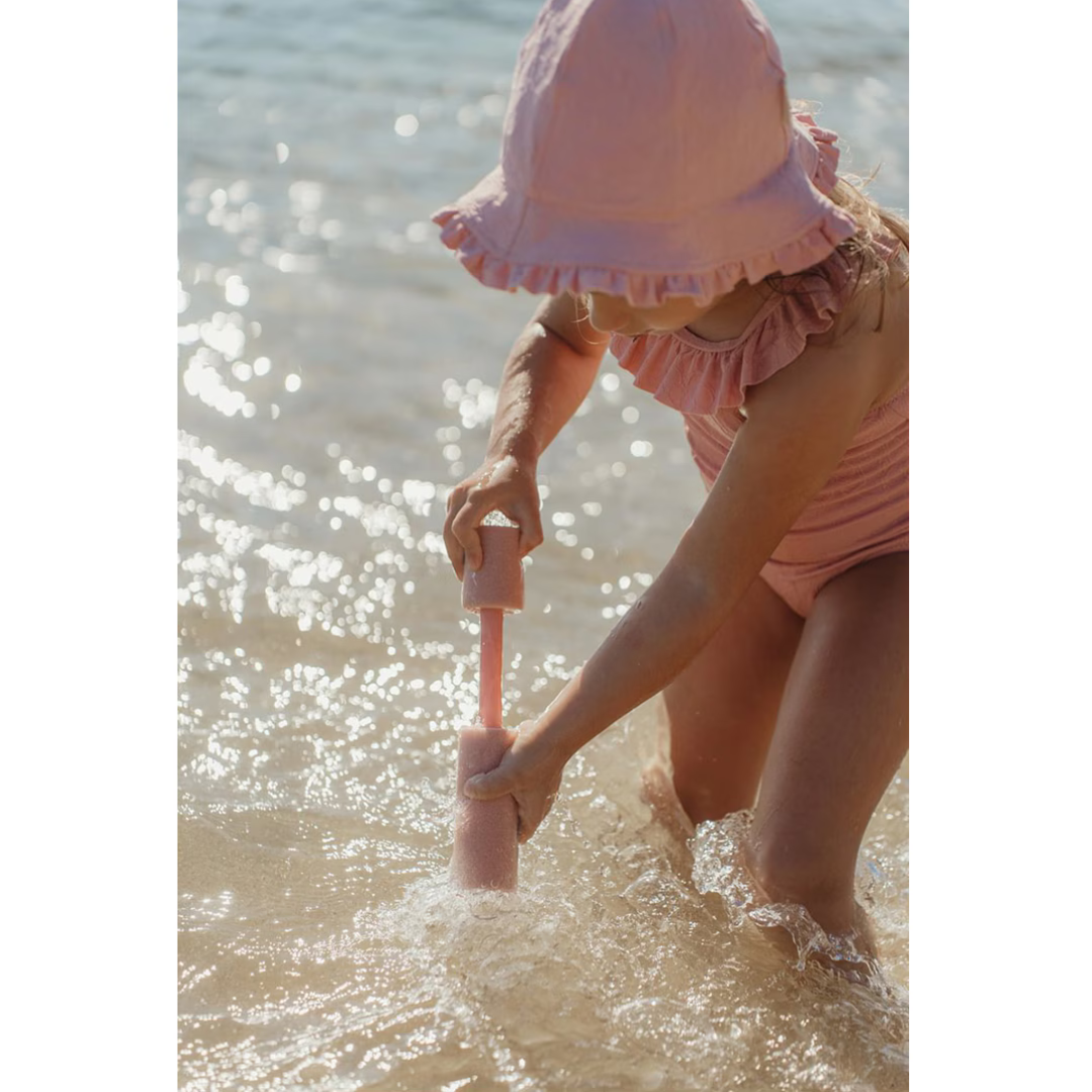 Menina de fato de banho e chapéu cor de rosa a brincar na água com ferramenta de areia cor de rosa.