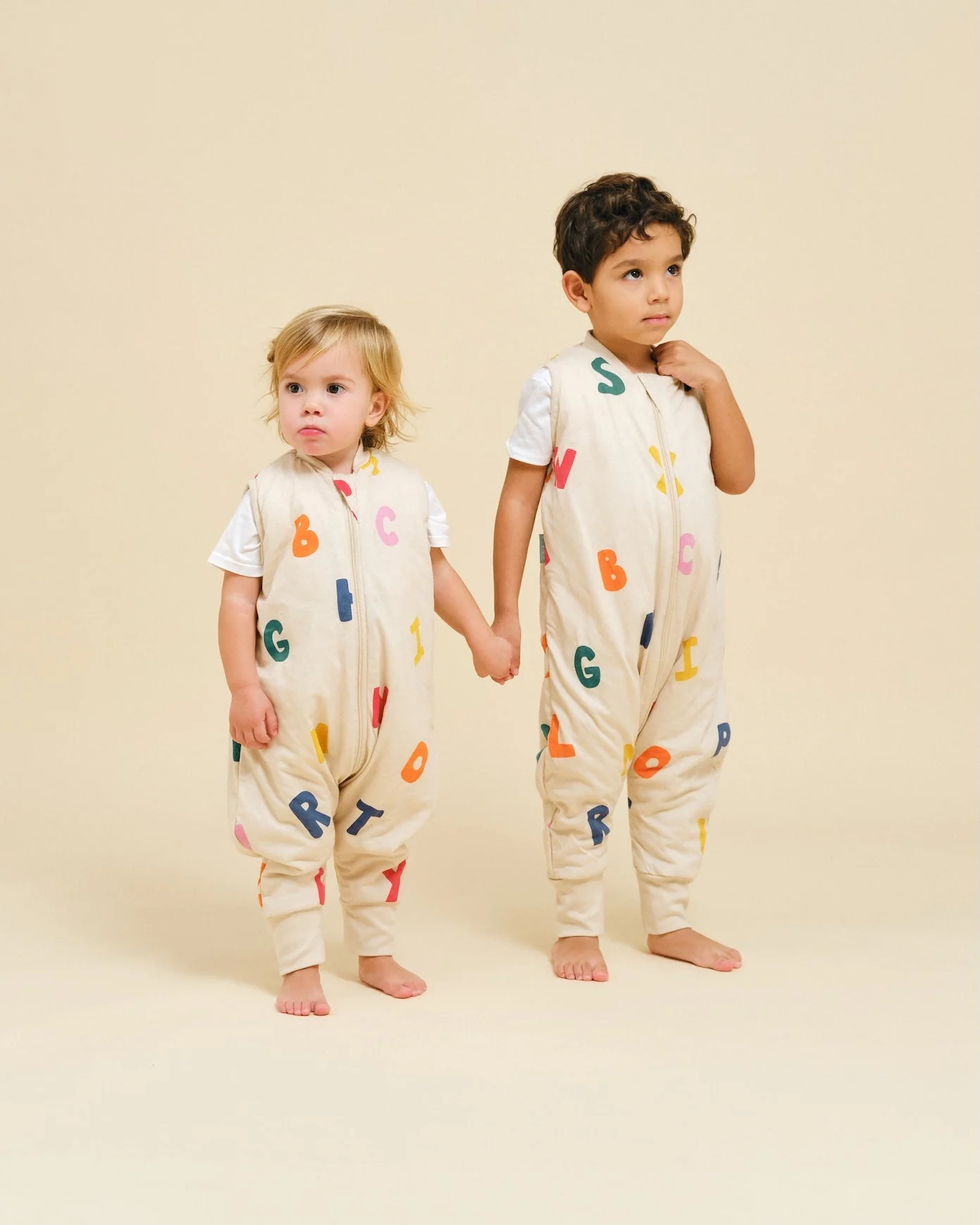 Duas crianças de baby grows creme com letras coloridas segurando as mãos