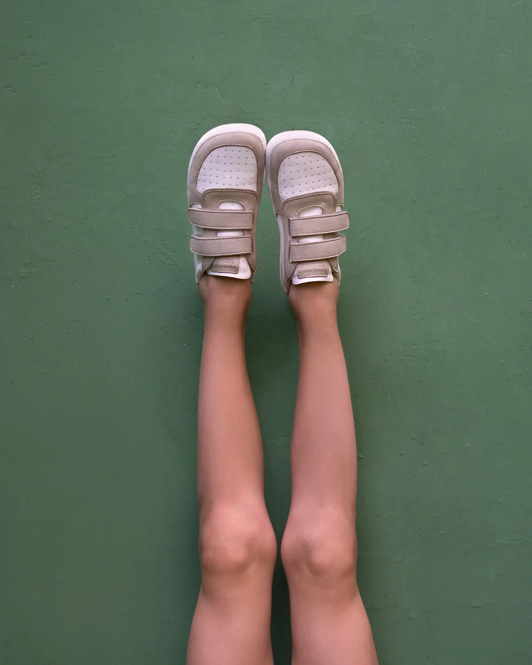 Sapatos desportivos infantis bege com fechos de velcro em pernas contra parede verde