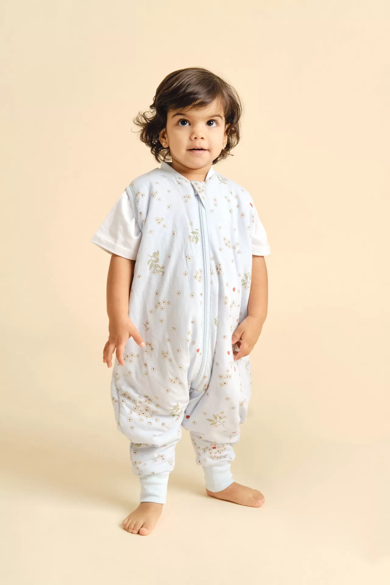 Criança usa babygrow azul claro com padrão floral e camisola branca em fundo bege