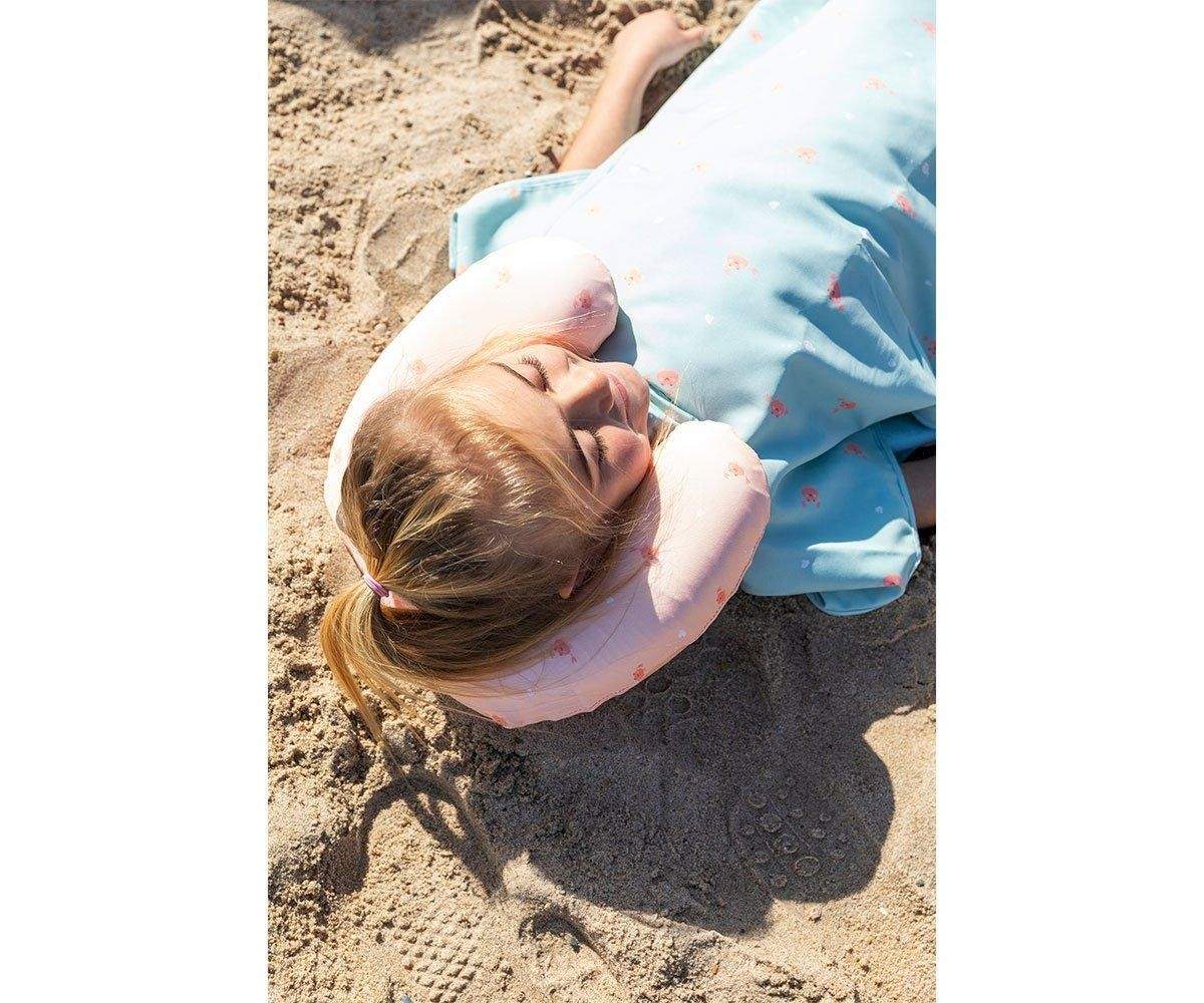 Criança deitada na areia usando vestido azul claro e almofada de pescoço inflável rosa claro