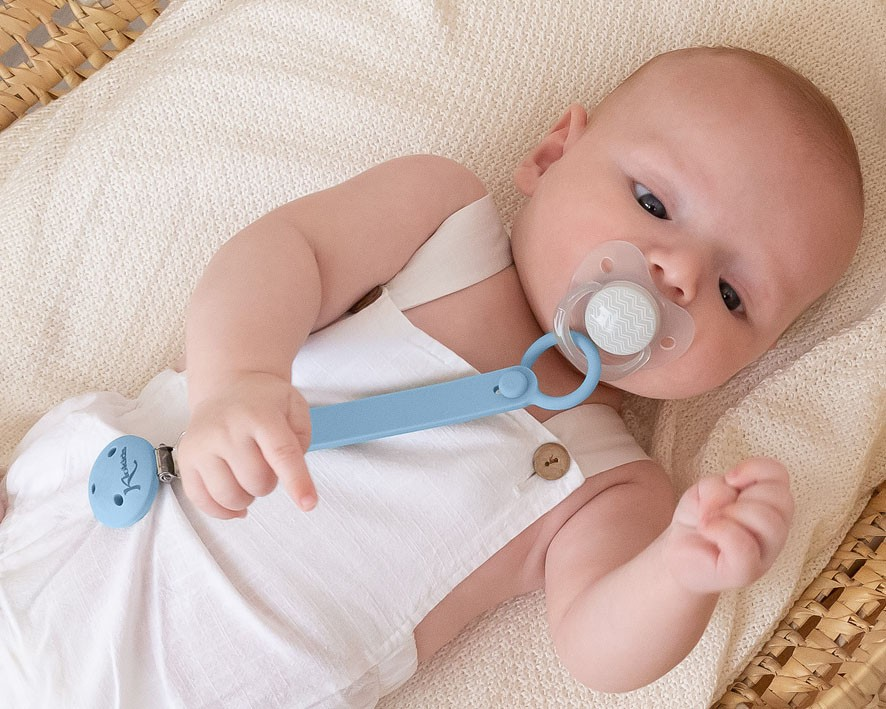 Bebé com chupeta cinzenta e clip azul numa almofada bege numa cesta de vime